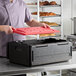 A black insulated food pan carrier with a red hot board lid being placed on top, designed for transporting and maintaining the temperature of food.
