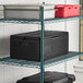 A black insulated food pan carrier with two full-size stainless steel food pans and lids, placed on a wire shelving unit.