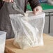 A person using a metal scoop to put flour into a clear gusseted polyethylene bag.