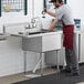 A man in a red apron washing his hands in a Regency stainless steel sink with a left drainboard.