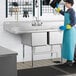 A person in a blue apron and yellow gloves washing dishes in a Regency stainless steel two compartment sink.