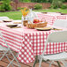 A picnic table with a red and white checkered tablecloth and a basket of fruit on a table set with white plates.