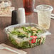 A salad in a clear plastic deli container with a lid on a table with a drink and white plastic utensils.