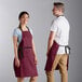 A man and woman wearing burgundy poly-cotton bib aprons with black webbing accents.