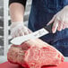 A person in gloves cutting raw meat on a counter with a Mercer Culinary American Butcher Knife.