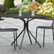 A black Lancaster Table & Seating outdoor table with two chairs on a stone patio.