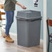 A woman standing next to a Lavex gray square trash can with a swing lid.