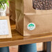 A brown paper bag with a clear window showing coffee beans and a round green and white Friday day-of-the-week clock label attached to the front.