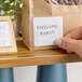 A removable blank label measuring 2 inches by 2 inches applied to a brown paper bag.