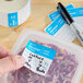 A removable blue and white Monday day-of-the-week label being applied to a food storage container, with a roll of similar labels and a marker nearby.