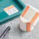 A roll of paper with orange and white Noble Products Dissolvable Day of the Week labels next to a pen.