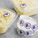 A roll of purple and white Point Plus Egg Allergen labels on a deli counter with plastic containers of food.