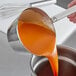 A person using a Choice stainless steel ladle to pour orange liquid into a pot.