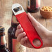 A hand using a Choice red bottle opener to open a brown bottle of beer.