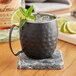 An Acopa hammered black Moscow Mule mug filled with ice and a lime wedge on a marble coaster.