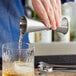 A hand using an Acopa stainless steel Japanese jigger to pour liquid into a glass on a metal bar.