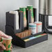 A hand holding a coffee cup and a Choice black metal countertop organizer with cups and condiments.