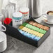 A Choice Black Countertop Coffee Condiment Organizer tray with condiments and sugar packets on a counter.