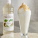 A glass of milkshake with whipped cream and coconut flakes in front of a bottle of Monin Premium Coconut Flavoring Syrup.