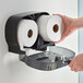 A hand using a Sierra Hygiene Little Big Roll twin dispenser to hold a roll of toilet paper.