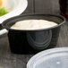 A close-up of a black Pactiv Ellipso oval souffle container with a clear lid filled with white sauce.