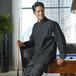 A man wearing a black Uncommon Chef Rio long sleeve chef coat leaning on a pool table.