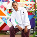 A man in a Uncommon Chef white long sleeve chef coat with black piping posing in front of a colorful mural.