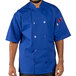 A man wearing a blue Uncommon Chef short sleeve chef coat with royal blue accents poses in a professional kitchen.