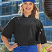 A woman wearing a black Uncommon Chef Aruba Pro Vent chef coat with a blue fabric knot in a professional kitchen.