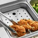 A stainless steel steam table pan with a false bottom on a counter with a tray of chicken and broccoli and tongs.