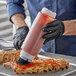 A person wearing black gloves using a Tablecraft INVERTAtop squeeze bottle to pour sauce over a piece of meat.