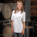 A woman wearing a Mercer Culinary white short sleeve brewer / work shirt standing in front of barrels.