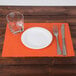 A Hoffmaster bittersweet orange paper placemat with scalloped edges under a plate and glass.