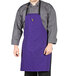 A man wearing a purple Uncommon Chef bib apron with black webbing standing in a professional kitchen.