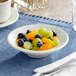 An Acopa ivory stoneware fruit bowl filled with oranges and grapes on a table with silverware.