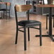 A Lancaster Table & Seating black wood chair with black vinyl seat and vintage wood back at a table in a restaurant.