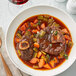 A white bowl of elk osso buco with vegetables and meat on a white plate.