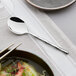 An Acopa Heika stainless steel bouillon spoon on a table next to a bowl of soup.