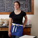 A woman wearing a black shirt and a royal blue waist apron with natural webbing standing in front of a chalkboard.