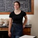 A woman wearing a navy blue waist apron with black webbing standing in front of a chalkboard.