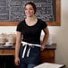 A woman wearing a Choice navy blue waist apron with natural webbing standing in front of a chalkboard.