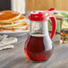 A Vollrath glass syrup dispenser with a red lid full of syrup on a table with pancakes.