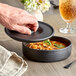 A hand using a black Tuxton tortilla server lid to cover a bowl of soup.