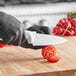 A person in black gloves using a Choice serrated chef knife to cut a tomato on a white counter.