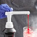 A gloved hand using a white Capora flavoring sauce pump to pour red liquid into a glass.