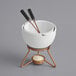 A white ceramic fondue pot with two copper forks on a stand over a candle.