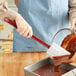A woman using a red Choice 16" heat-resistant silicone spatula to mix liquid chocolate in a pan on a counter.