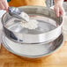 A person pouring flour into a Choice stainless steel sieve.