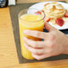 A hand holding a Cambro Newport plastic tumbler filled with orange juice on a table in a breakfast diner.
