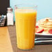 A Cambro clear plastic tumbler filled with orange juice on a table with pancakes.